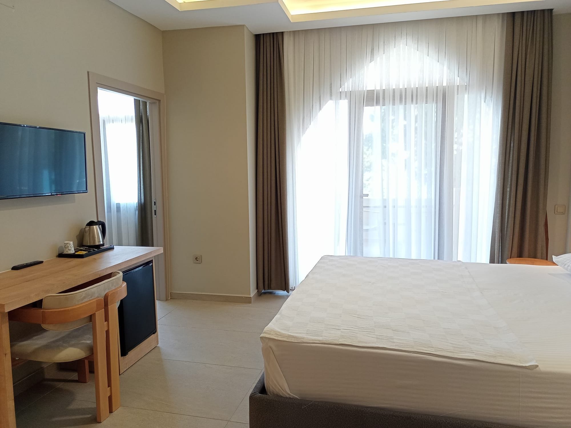 A hotel room with a bed, a desk with a television, and a window with curtains.
