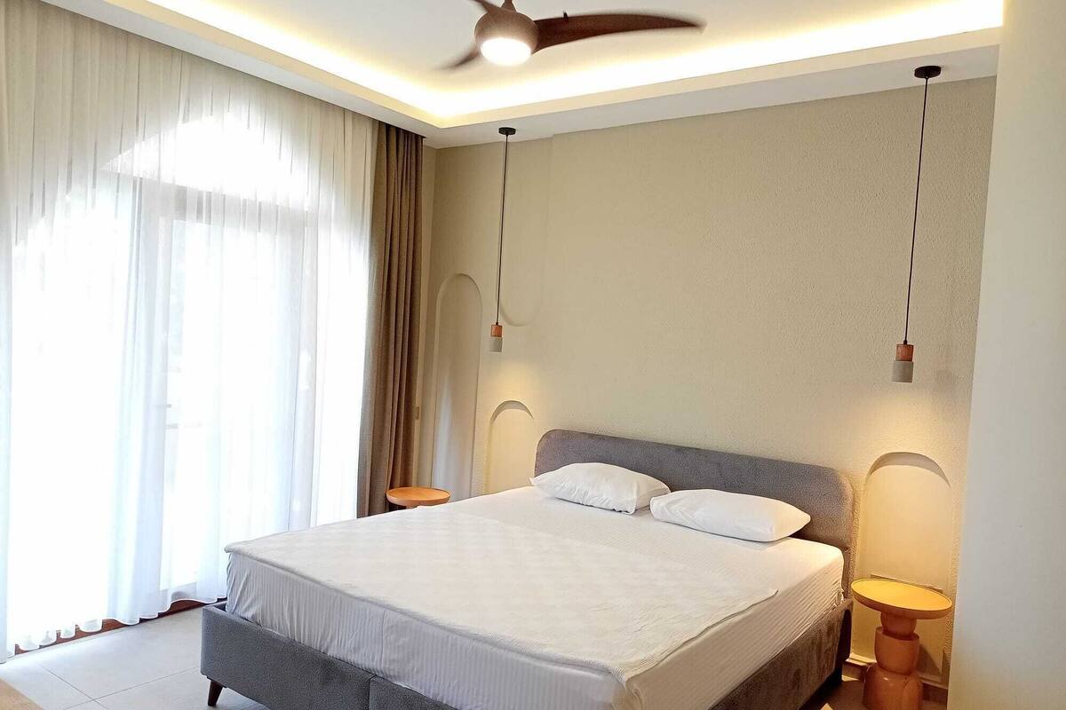 Een moderne hotelkamer met een groot bed, twee nachtkastjes, een plafondventilator en een groot raam met gordijnen.