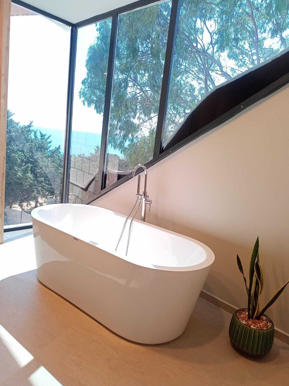 A modern bathroom with a freestanding bathtub, large windows, and a view of trees and a body of water.