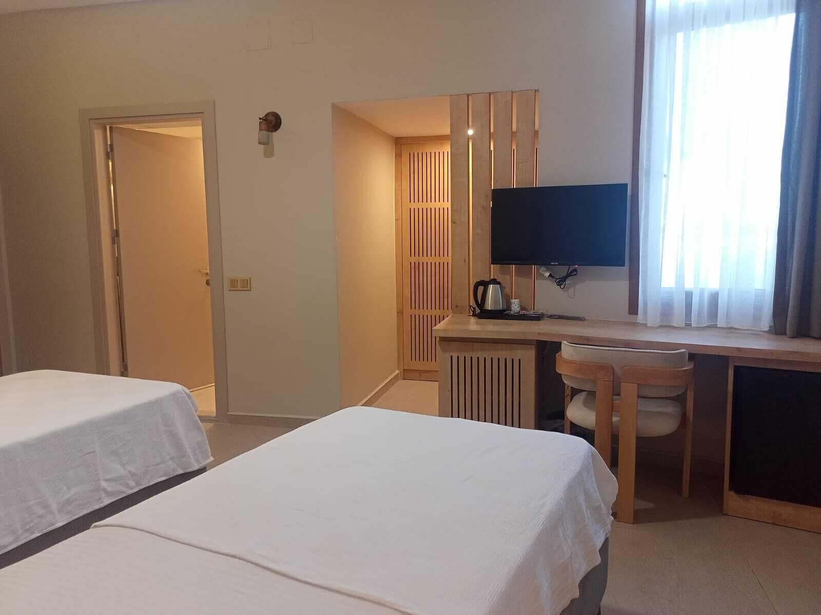 A hotel room with two single beds, a desk with a chair, a TV, and a window with curtains.