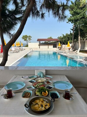 A poolside dining area with a table set for breakfast, overlooking a swimming pool and a building with a red roof. - Casalita Hotel (Erdemli)