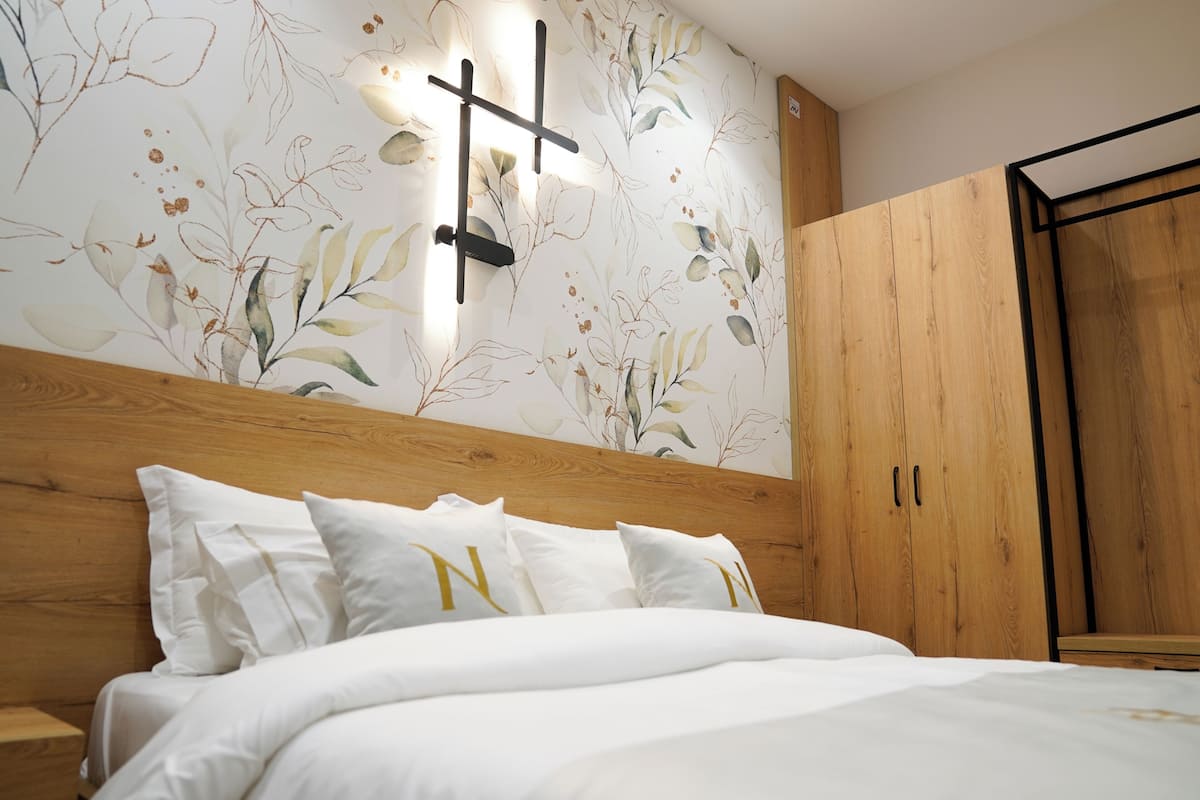 A bedroom with a wooden headboard, a bed with white bedding, a wall-mounted light fixture, and a wooden wardrobe.