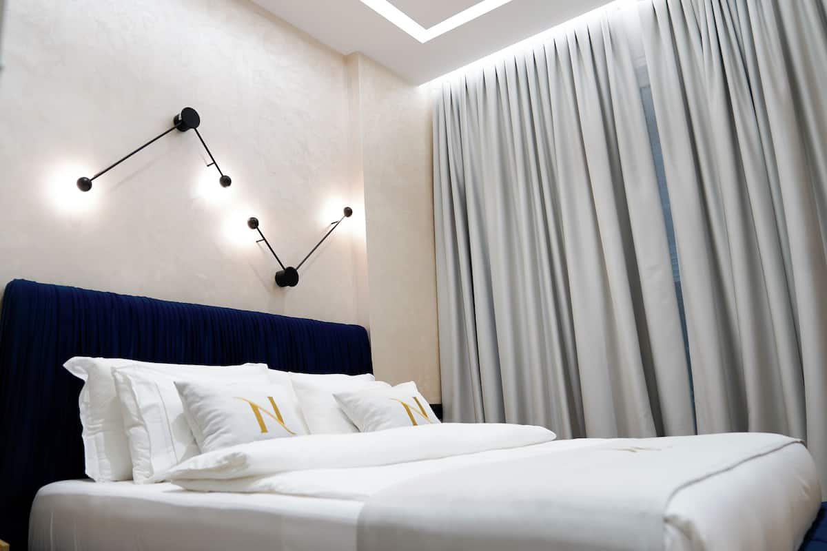 A modern hotel room with a neatly made bed, a wall-mounted light fixture, and a large window with curtains.