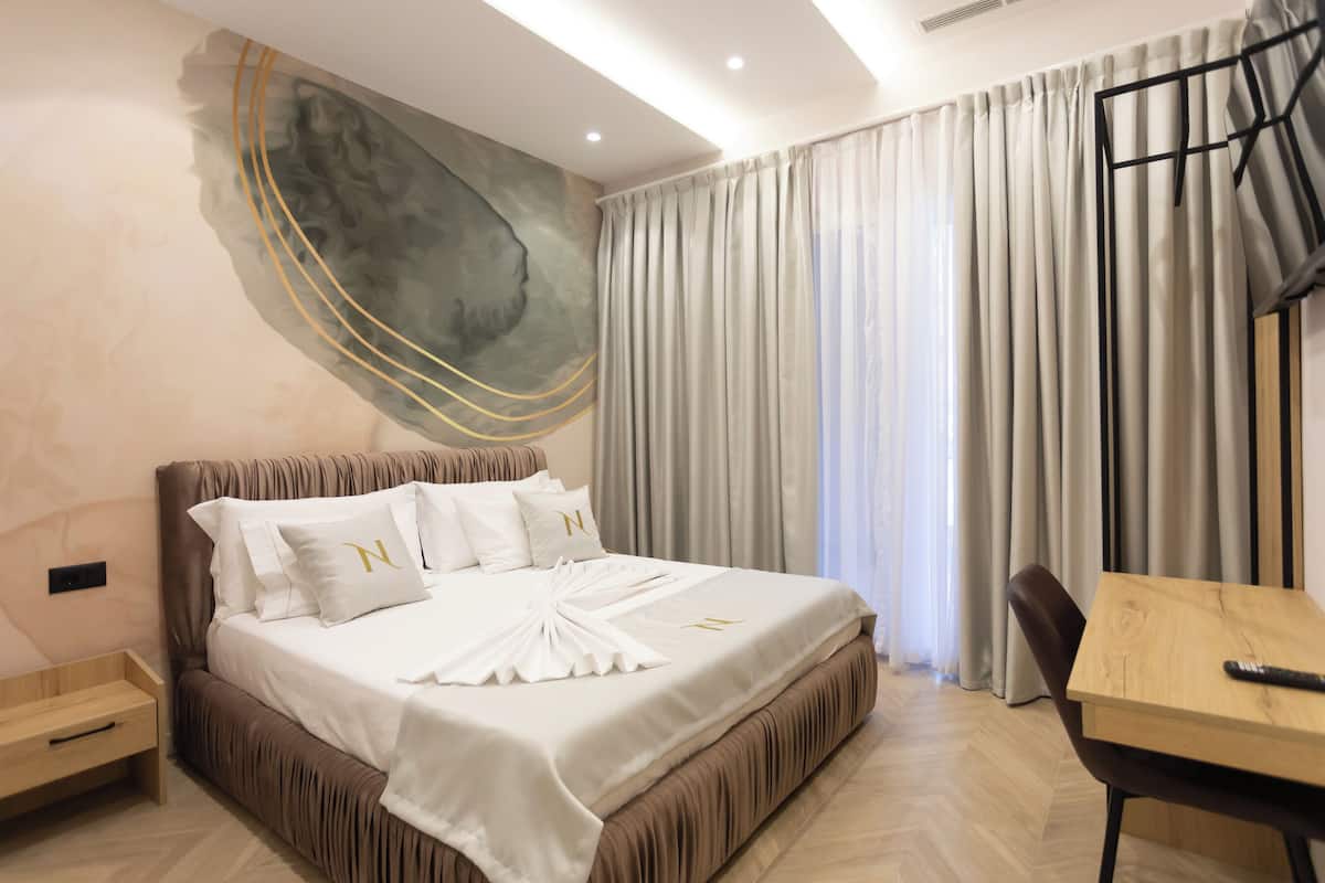 A modern hotel room with a large bed, a wooden table, and a view of the city through sheer curtains.