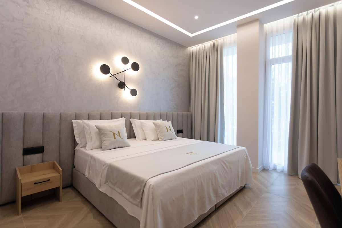 A modern bedroom with a large bed, a bedside table, a wall-mounted light fixture, and a view of the outdoors through large windows.