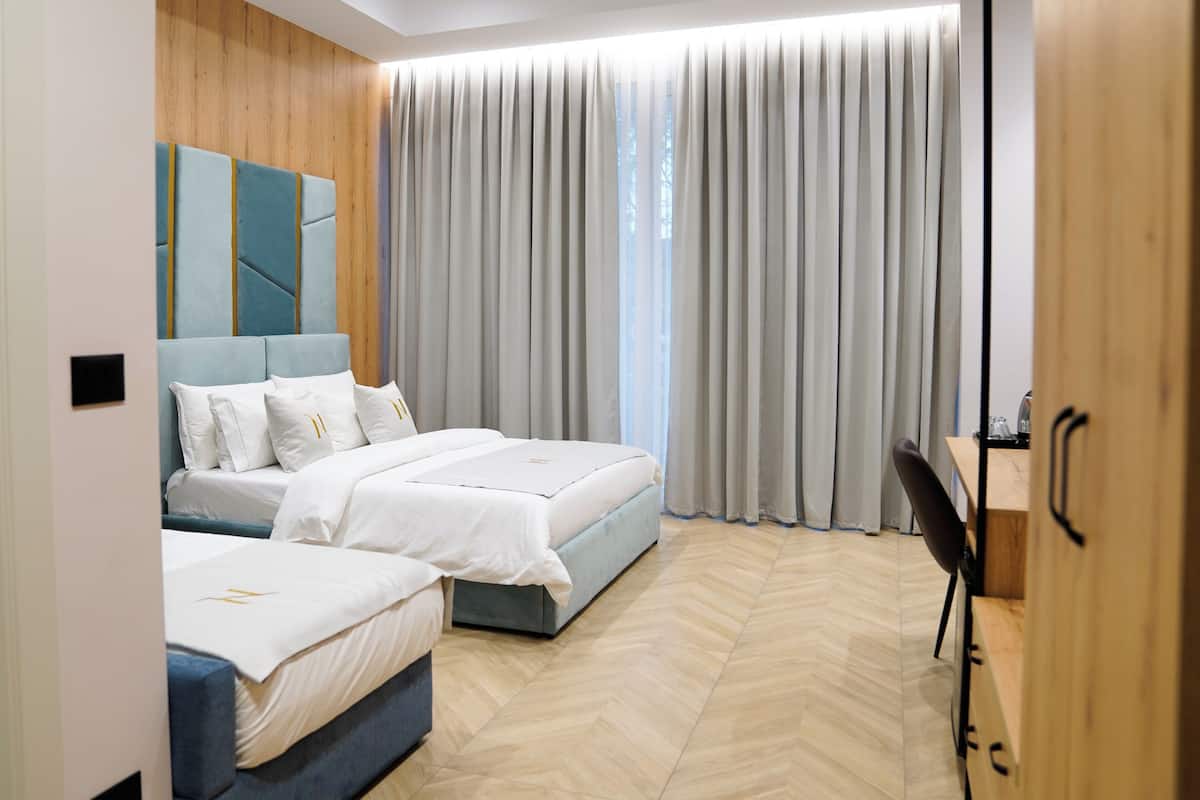 A modern hotel room with two beds, a wooden accent wall, a large window with curtains, and a desk with a chair.