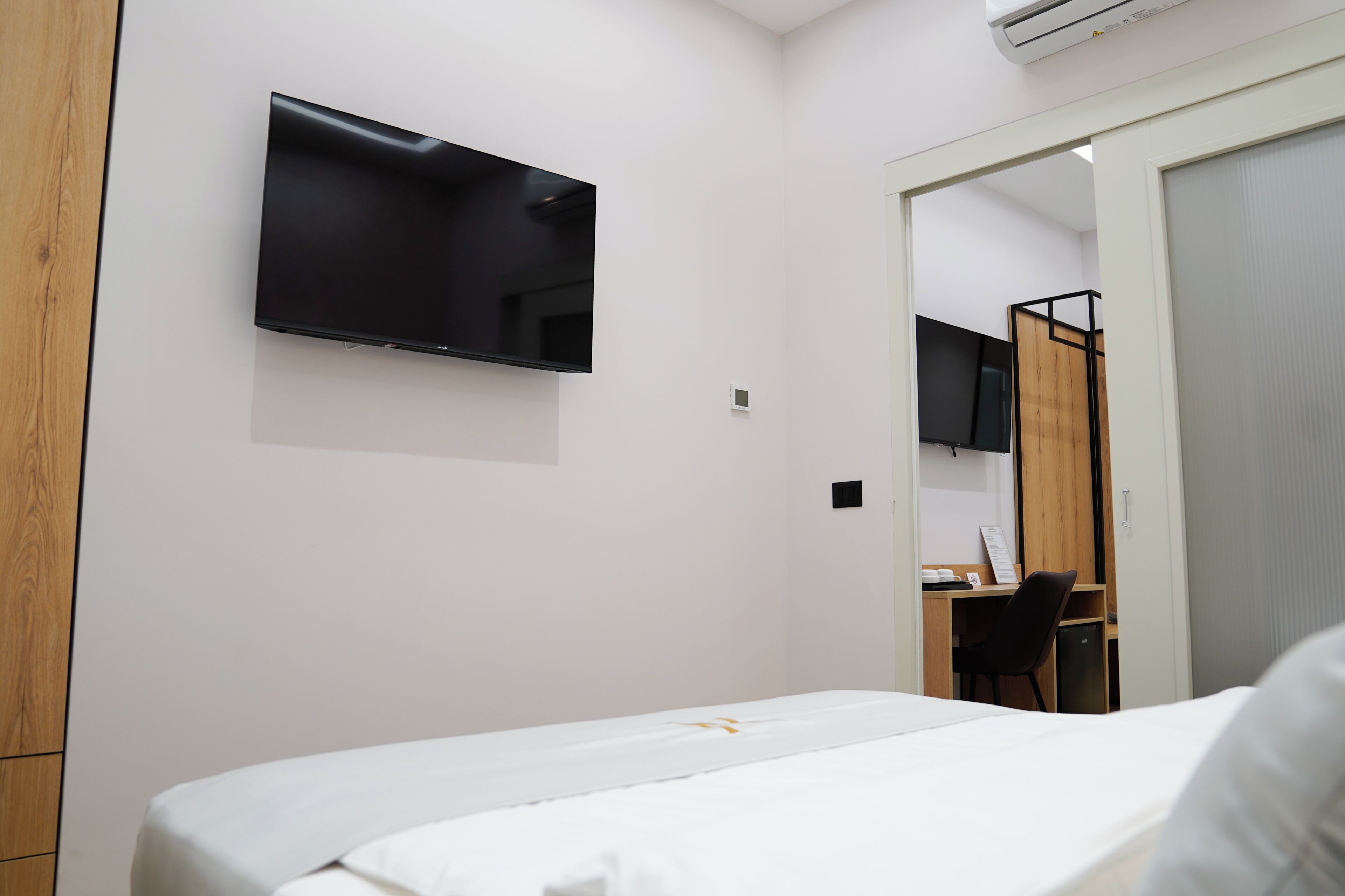 A hotel room with a bed, a desk, a chair, a TV mounted on the wall, and a door leading to another room.