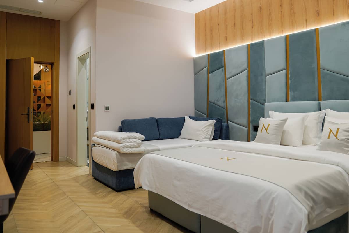A modern hotel room with a large bed, a smaller sofa, a wooden headboard, and a dining area.