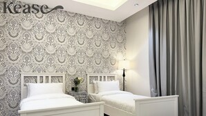 Une chambre avec deux lits blancs, un papier peint à motifs, une table de chevet avec une lampe et un rideau gris.