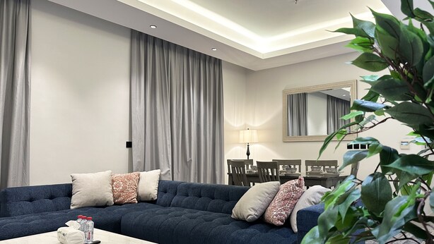 Un salon moderne avec un canapé bleu foncé, des rideaux gris, une table à manger avec des chaises et un grand miroir au mur.