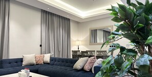 A modern living room with a dark blue sofa, grey curtains, a dining table with chairs, and a large mirror on the wall. - Kease Al Wahah K-2 Royal Touch AX68 (Riyadh)