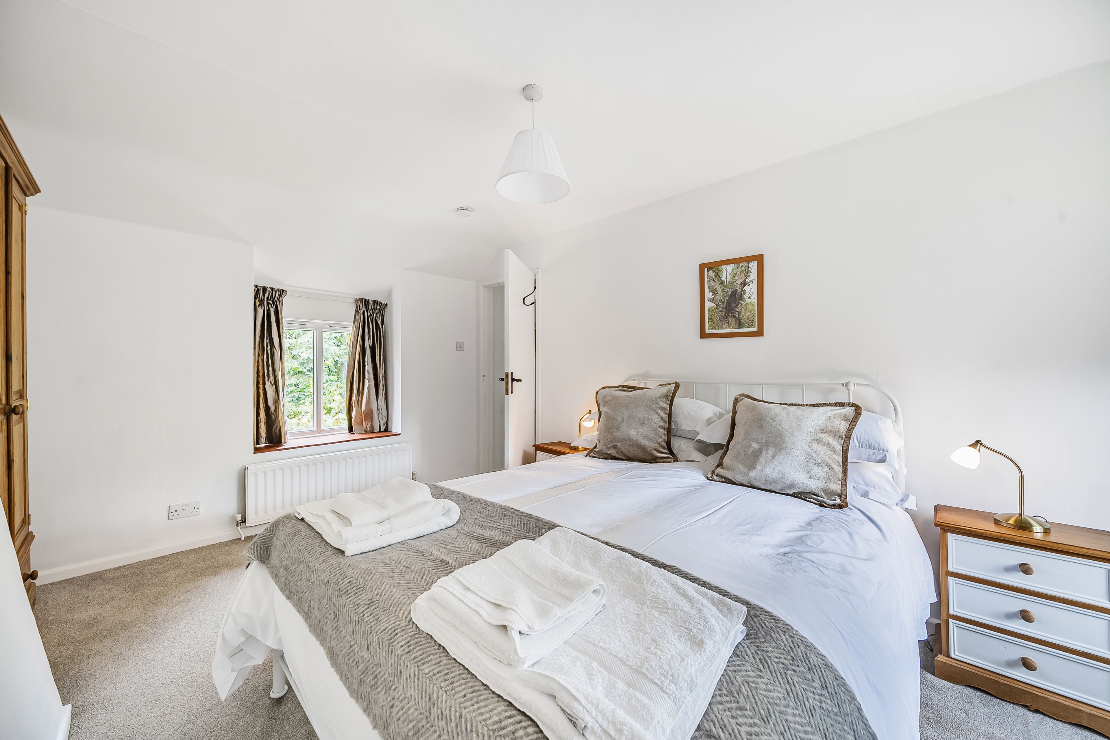 Une chambre à coucher bien rangée, avec un lit, des tables de chevet, une commode, une fenêtre avec des rideaux et un radiateur.