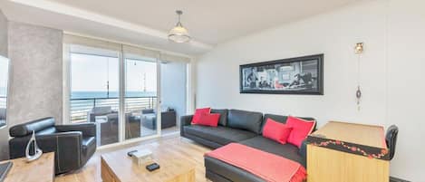 A modern living room with a sofa, coffee table, and a desk with a lamp. There is a large window with a view of the sea.