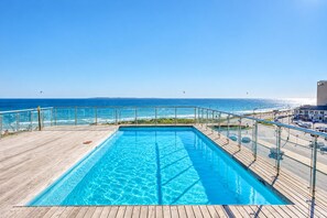 A rectangular swimming pool with a wooden deck and glass railing, overlooking a beach and ocean.