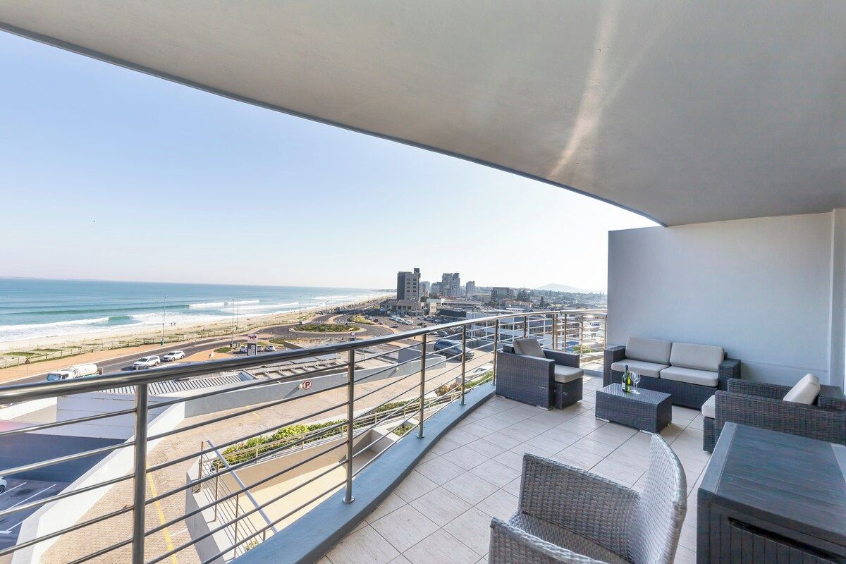 A balcony with a view of the beach and cityscape, furnished with outdoor seating and a railing.