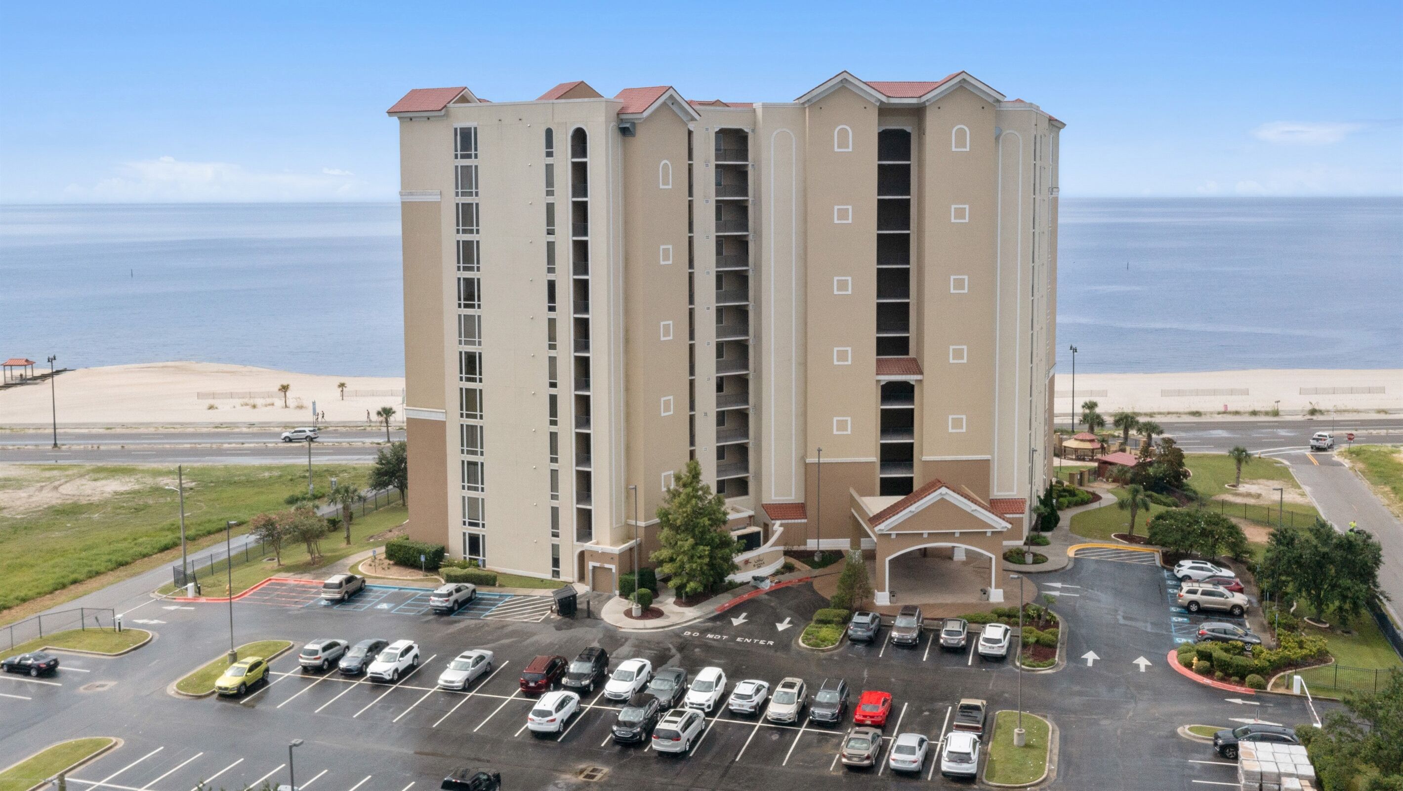 A multi-story hotel with a parking lot in front, located near a beach.
