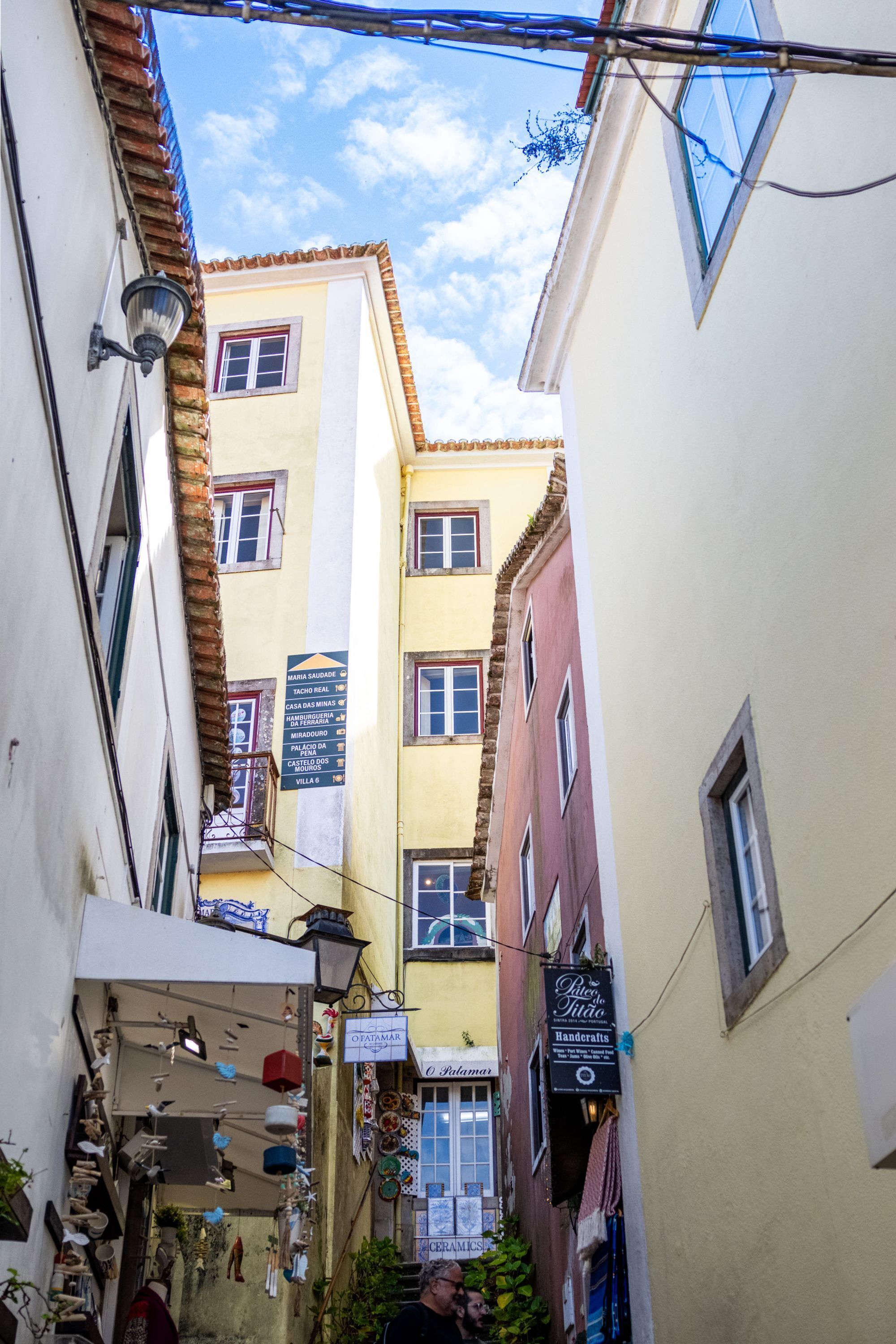 A narrow street with buildings on either side, featuring balconies and a variety of shops.