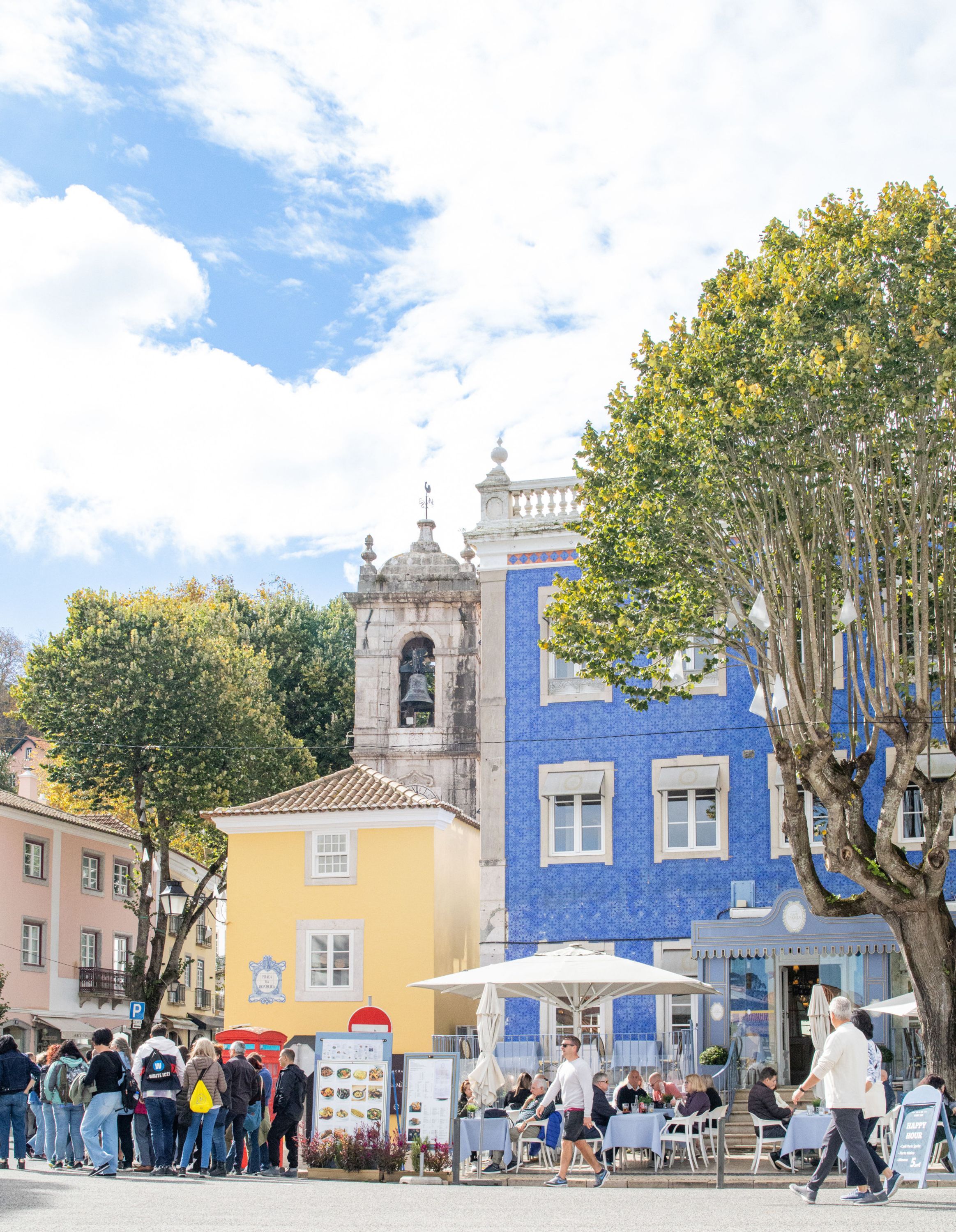 A public square with a historic bell tower, a blue building with tiled walls, and a tree.