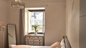 A bedroom with a bed, a dresser, a mirror, a chandelier, and a window with a view of buildings. - Sintra Historic Centre Romantism Capital (Sintra)