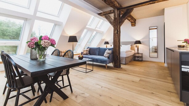 A modern loft bedroom with a sloped ceiling, skylights, a dining area with a wooden table and chairs, a blue sofa, and a kitchen area. - Gut Damp (Damp)
