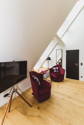 A modern loft bedroom with a sloped ceiling, a flat-screen TV, two red velvet chairs, a wooden floor, and a black door. - Gut Damp (Damp)