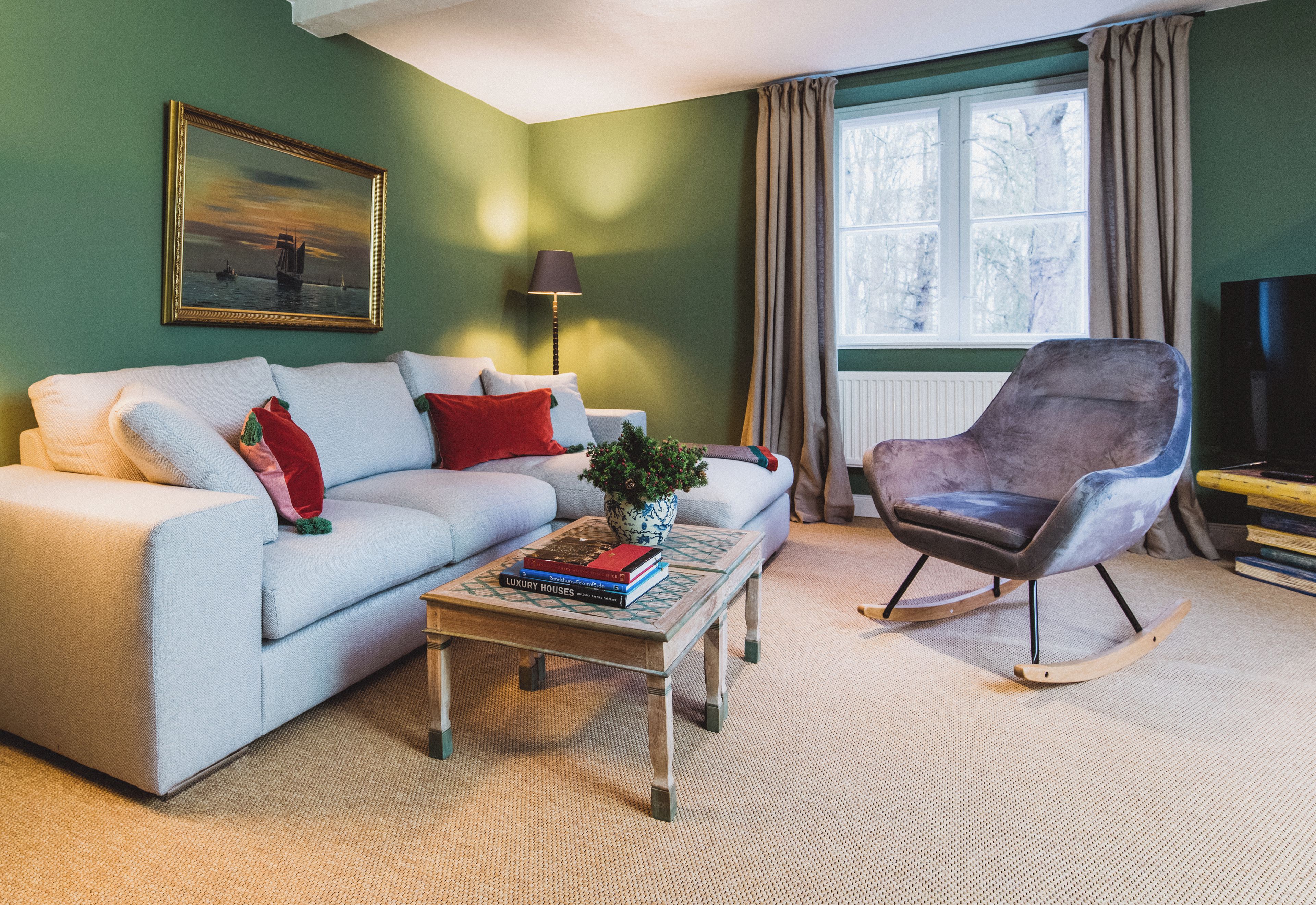 Een woonkamer met een bank, een schommelstoel, een salontafel met boeken, een lamp en een raam met gordijnen.