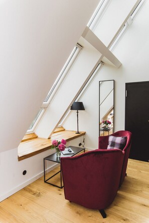 A modern loft bedroom with a sloped ceiling, a wooden desk, a red chair, a small table with a vase of flowers, and a black lamp.