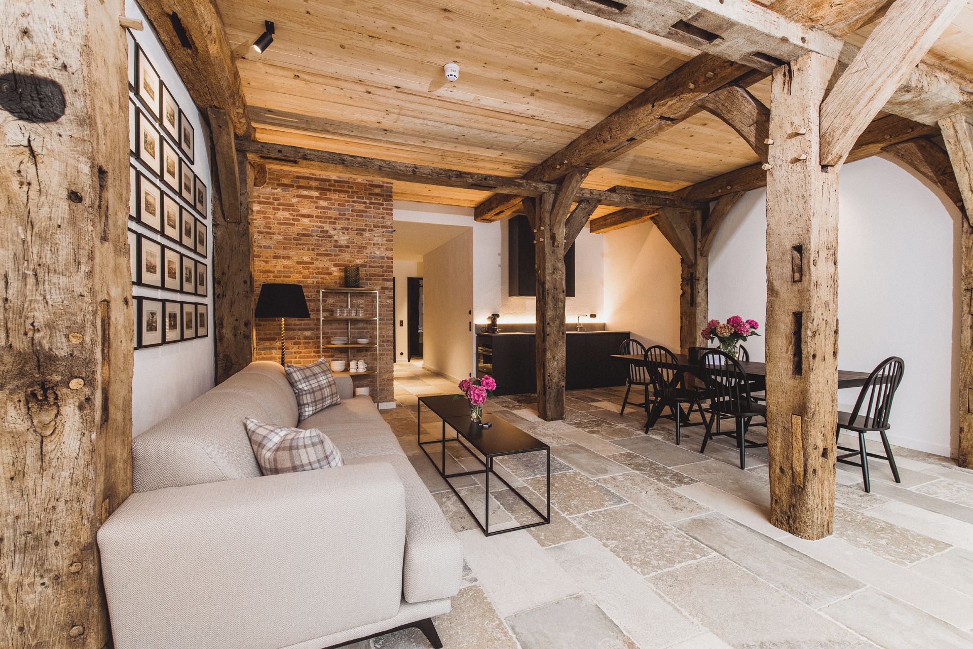 A rustic interior with exposed wooden beams, a stone floor, a sofa, a dining table with chairs, and a kitchen area.