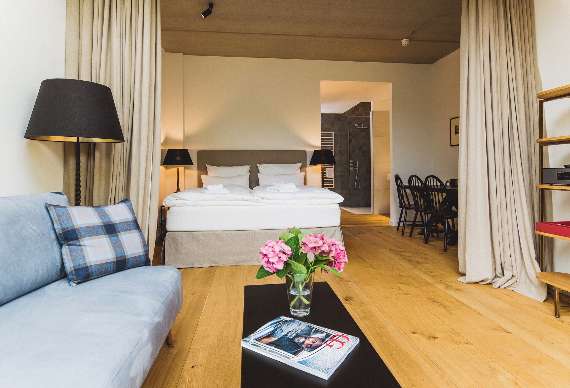 A hotel room with a bed, a sofa, a dining table, and a coffee table with a magazine and flowers.