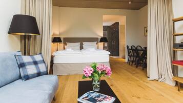 A hotel room with a bed, a sofa, a dining table, and a coffee table with a magazine and flowers.