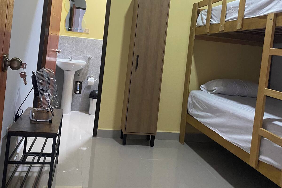A compact room with a bunk bed, a wooden wardrobe, a small table with a fan, and a bathroom visible through an open door.