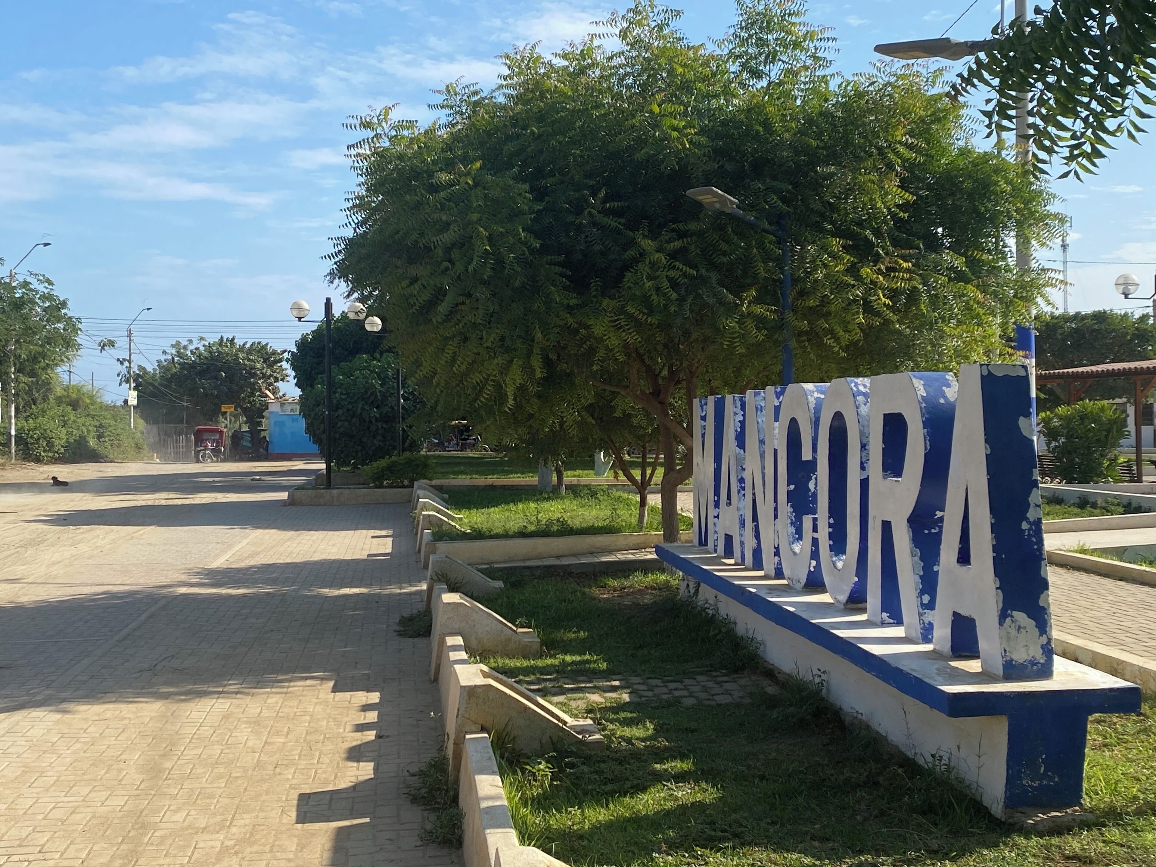 Un letrero con la palabra 'MANCORA' en letras grandes, rodeado de follaje y una zona pavimentada.