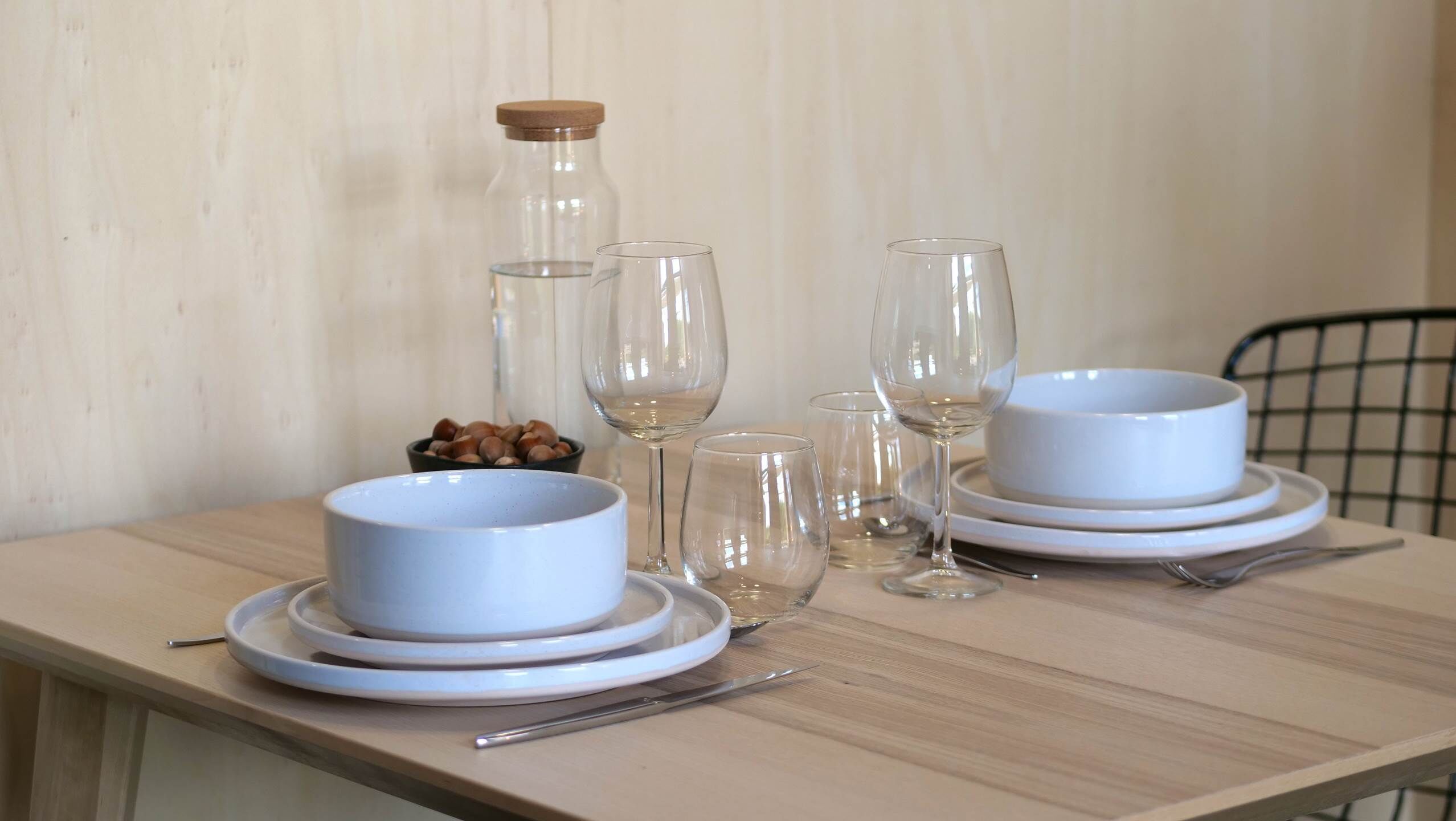 Une table à manger dressée avec des bols, des assiettes et des verres à vin blancs, ainsi qu’une bouteille et un bol de noix.
