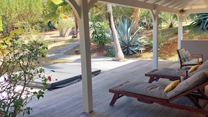 A covered outdoor area with wooden deck, lounge chairs, and a small seating area with cushions. - Jardin d'O résidence naturiste couples only (Orient Bay)
