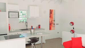 A modern kitchen with a microwave, sink, and a window with a view of greenery. There is a dining area with a table and chairs, and a red sofa with cushions. - Jardin d'O résidence naturiste couples only (Orient Bay)