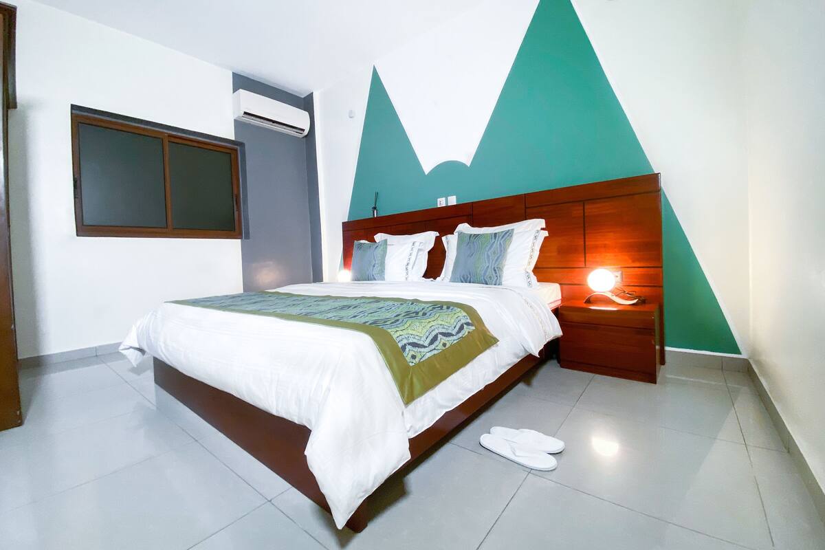 A hotel room with a bed, a nightstand with a lamp, a window, and an air conditioning unit.