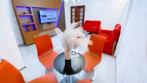 Ein modernes Wohnzimmer mit Glastisch, orangefarbenen Sesseln, einem roten Sofa, einem Fernseher auf einem Holzregal und einer Deckenleuchte.