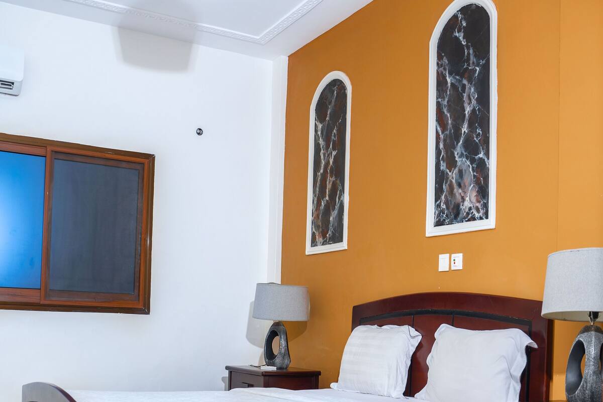 A bedroom with a wooden bed, two bedside tables with lamps, a large window, and a wall-mounted air conditioner.