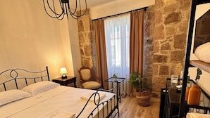A bedroom with a bed, a chandelier, a stone wall, a window with curtains, a chair, a table, and a plant. - Milestone Otel (Ayvalik)
