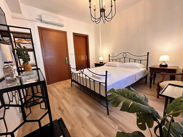 A bedroom with a bed, bedside tables, a desk, and a chair. There is a chandelier, a plant, and an air conditioning unit.