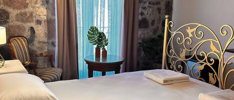 Una habitación acogedora con pared de piedra, vigas de madera en el techo, cama con ropa blanca, cabecero de metal con diseños de animales, una mesita con una planta y una ventana con cortinas.