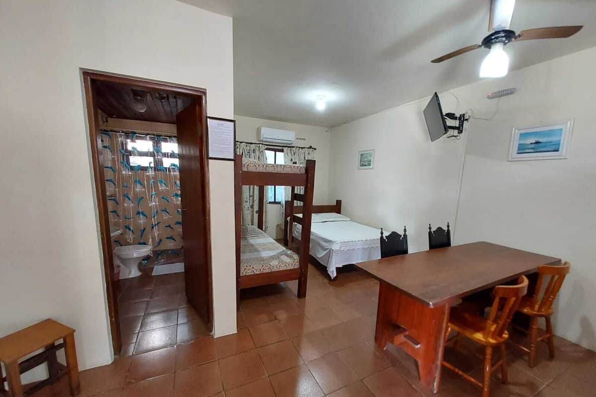 Ein Schlafsaalzimmer mit Etagenbett, einem kleinen Tisch und Stühlen. Es gibt einen Deckenventilator, eine Klimaanlage und ein Fenster mit Vorhängen.