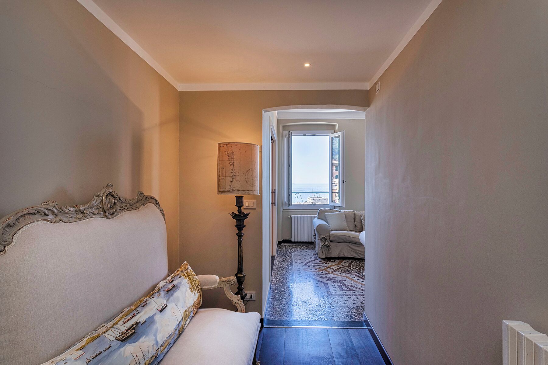 A narrow hallway with a bed on the left, a lamp, and a view of a room with a window and a chair.