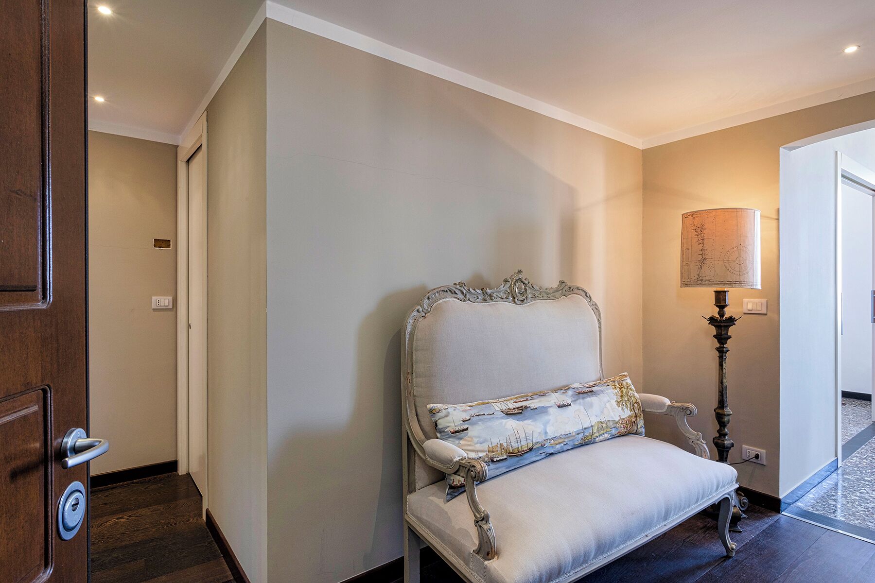 Un couloir avec une porte en bois, un fauteuil blanc recouvert de tissu avec un coussin décoratif et un lampadaire.