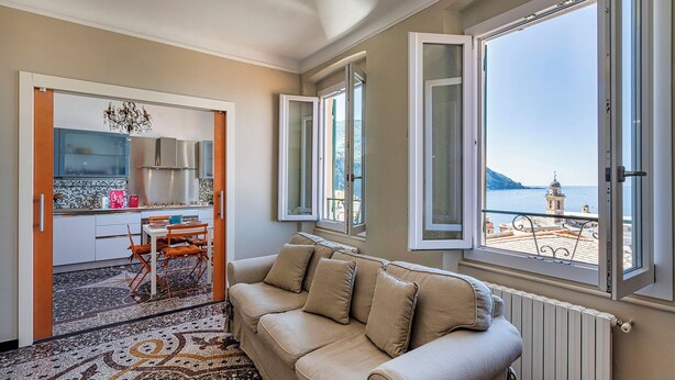 A cozy living room with a beige sofa, a dining area with a table and chairs, and large windows offering a view of the sea and a bell tower. - Camogli Gulf View by Wonderful Italy (Camogli)