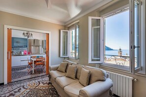 Ein gemütliches Wohnzimmer mit einem beigen Sofa, einem Essbereich mit Tisch und Stühlen und großen Fenstern mit Blick auf das Meer und einen Kirchturm.