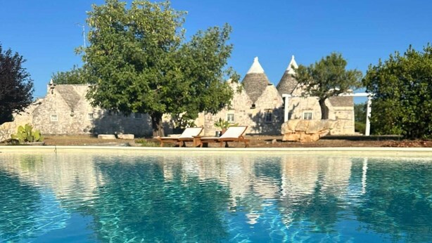 A pool with clear water, surrounded by stone buildings with conical roofs, and two lounge chairs by the poolside. - Trulletto with swimming pool in Monopoli (Monopoli)