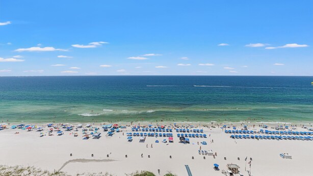Una playa con numerosas sombrillas y tumbonas, cielo despejado y mar tranquilo.