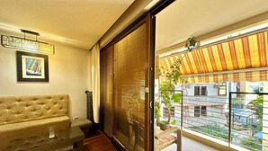 A living room with a sofa, a glass coffee table, and a sliding glass door leading to a balcony with a view of a building and a plant. - Family Two Bedroom Apartment Vasko (Budva)