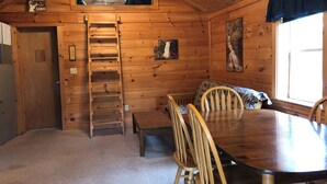 Interior de una cabaña con una mesa de comedor de madera, sillas, una escalera, un banco y una ventana con vistas.